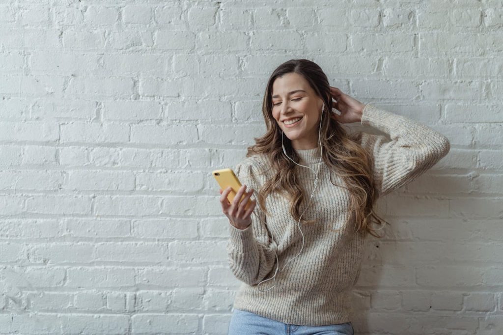 Smiling young woman using smartphone with earphones against a white brick wall.
